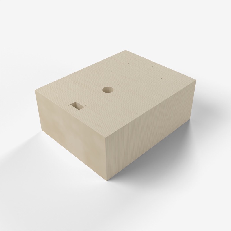 Three-quarter view of the base: a pale birch block, wider than it is tall, with a round hole, a square opening and small pin holes in its top face.